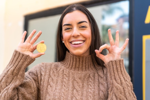 Jeune femme tenant un bitcoin à l'extérieur montrant un signe ok avec les doigts