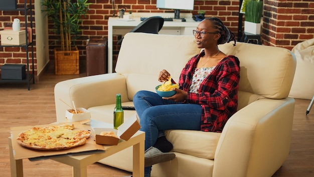 Jeune femme tenant un bol de chips en regardant la télévision, en buvant de la bière et en mangeant un repas de livraison à partir de plusieurs commandes de restauration rapide. Femme adulte ayant des hamburgers, des frites, des pizzas et des nouilles sur la table.