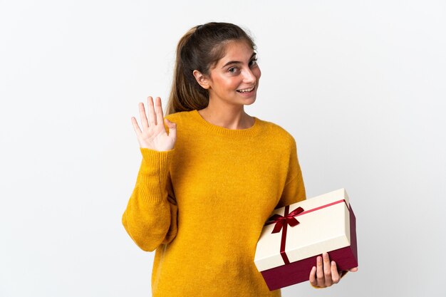 Jeune femme tenant un cadeau isolé sur blanc saluant avec la main avec une expression heureuse