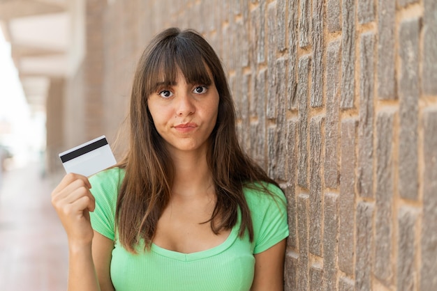 Jeune femme tenant une carte de crédit à l'extérieur avec une expression triste