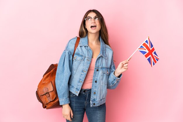 Jeune femme tenant un drapeau du Royaume-Uni isolé sur rose en levant et avec une expression de surprise