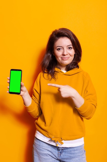 Jeune femme tenant un écran vert vertical sur le téléphone. Personne de race blanche pointant vers le modèle de maquette isolé et l'arrière-plan avec clé chroma sur smartphone. Adulte avec téléphone portable sur caméra