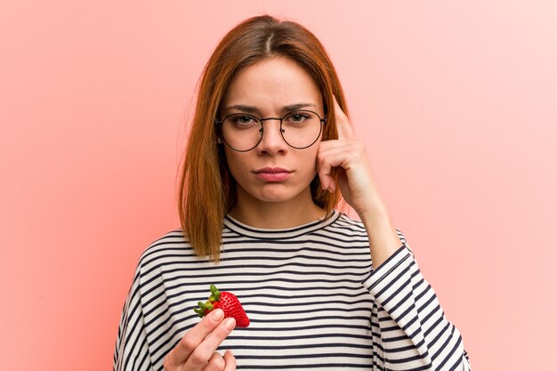 Jeune femme tenant une fraise en pointant sa tempe avec le doigt, pensant, concentrée sur une tâche.