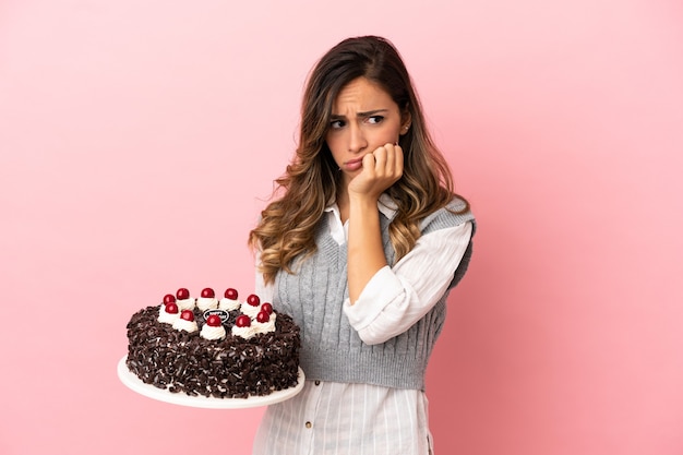 Jeune femme tenant un gâteau d'anniversaire sur fond rose isolé ayant des doutes