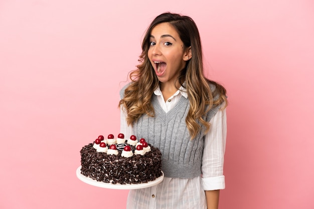 Jeune femme tenant un gâteau d'anniversaire sur fond rose isolé avec une expression faciale surprise