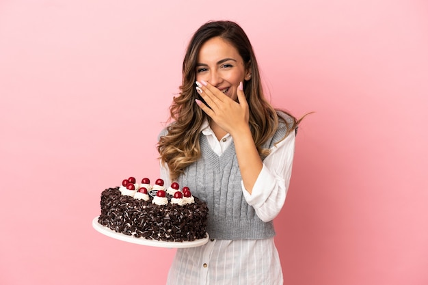 Jeune femme tenant un gâteau d'anniversaire sur fond rose isolé heureux et souriant couvrant la bouche avec la main