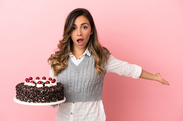 Jeune femme tenant un gâteau d'anniversaire sur fond rose isolé tendant les mains sur le côté pour inviter à venir