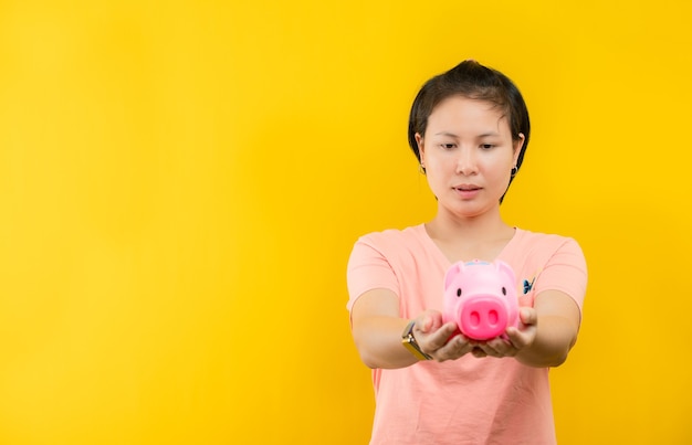 Jeune femme tenant joyeusement une tirelire rose pour collecter de l'argent s'affiche sur un fond jaune.concept d'épargne.