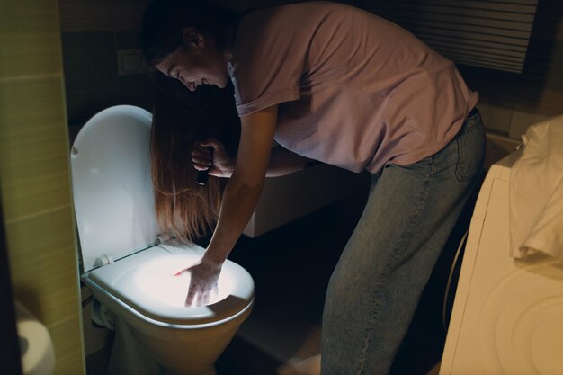 Une jeune femme tenant une lampe de poche dans l'obscurité regarde dans les toilettes.