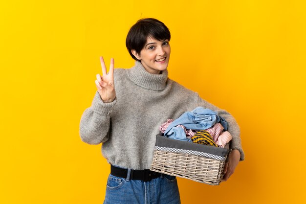 Jeune femme tenant un panier de vêtements souriant et montrant le signe de la victoire