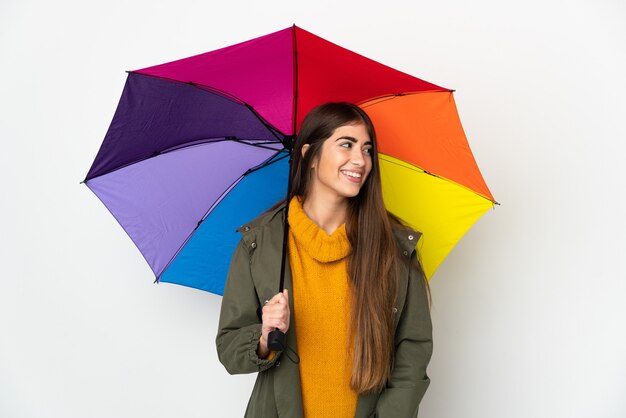 Jeune femme tenant un parapluie isolé sur un mur blanc à la recherche sur le côté et souriant