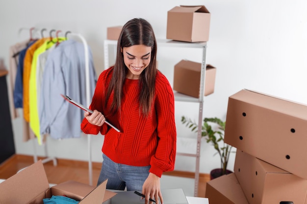 Jeune femme tenant le presse-papiers, regardant un ordinateur portable pour l'adresse du client. Propriétaire de petite entreprise, vente en ligne, boîtes d'emballage et livraison