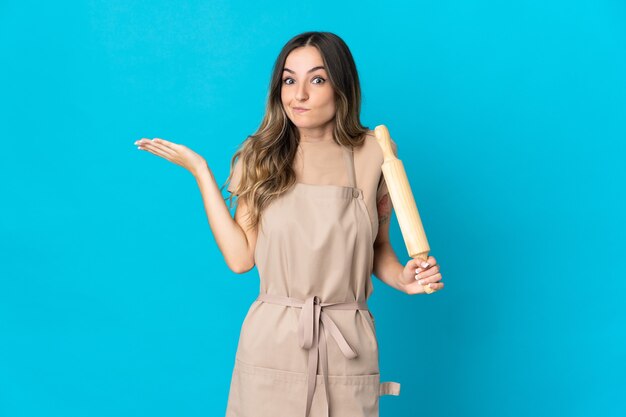 Jeune femme tenant un rouleau à pâtisserie isolé ayant des doutes tout en levant les mains