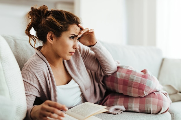 Jeune femme tenant sa tête dans la douleur en position allongée sur le canapé et lisant un livre à la maison