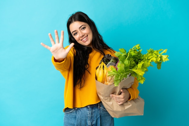 Jeune femme tenant un sac d'épicerie sur bleu en comptant cinq avec les doigts