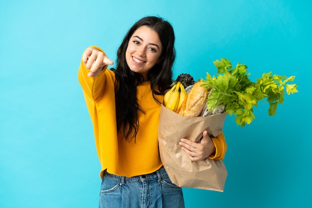 Jeune femme tenant un sac d'épicerie isolé sur le devant de pointage bleu avec une expression heureuse