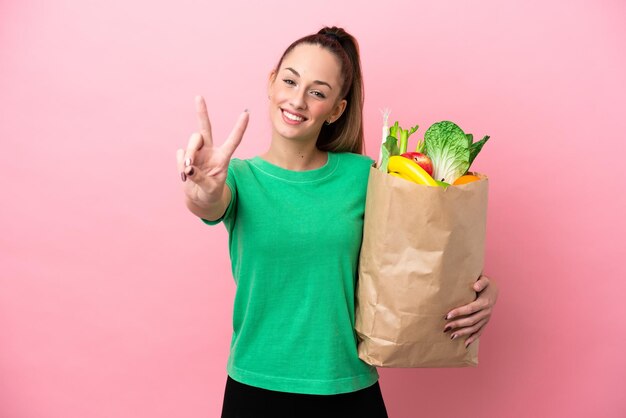 Jeune femme tenant un sac d'épicerie souriant et montrant le signe de la victoire