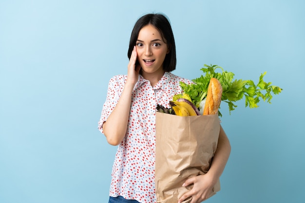 Jeune femme tenant un sac d'épicerie avec surprise et expression faciale choquée