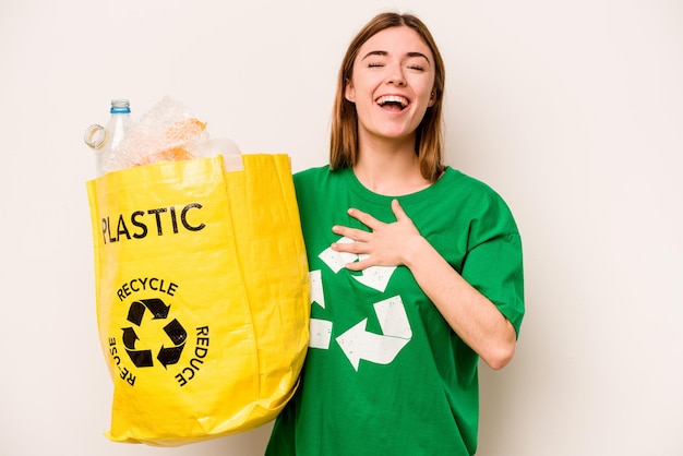 Jeune femme tenant un sac plein de bouteilles en plastique à recycler isolé sur fond blanc éclate de rire en gardant la main sur la poitrine