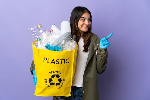 Jeune femme tenant un sac plein de bouteilles en plastique à recycler isolé sur fond violet pointant vers le haut une excellente idée