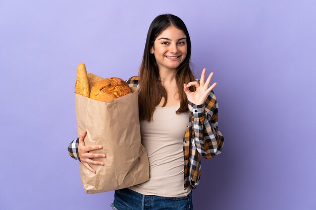 Jeune femme tenant un sac plein de pains isolé sur violet montrant signe ok avec les doigts