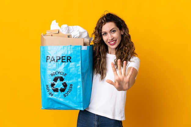 Jeune femme tenant un sac de recyclage plein de papier à recycler isolé sur jaune en comptant cinq avec les doigts