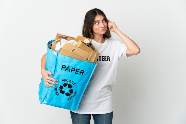 Jeune femme tenant un sac de recyclage plein de papier à recycler isolé sur un mur blanc ayant des doutes et de la réflexion