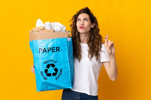 Jeune femme tenant un sac de recyclage plein de papier à recycler isolé sur mur jaune avec les doigts qui se croisent et souhaitant le meilleur