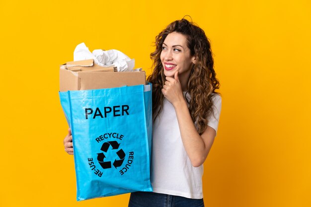 Jeune femme tenant un sac de recyclage plein de papier à recycler isolé sur mur jaune à la recherche sur le côté et souriant