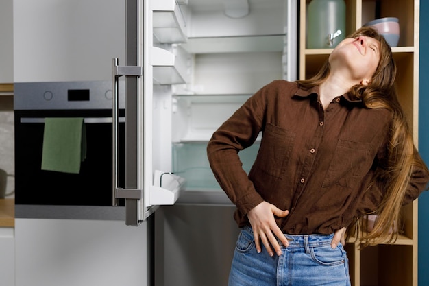 Jeune femme tenant son estomac debout près du réfrigérateur dans la cuisine