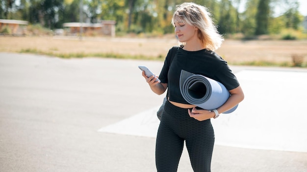 Jeune femme tenant un tapis de yoga et utilisant un smartphone après l'entraînement dans la rue