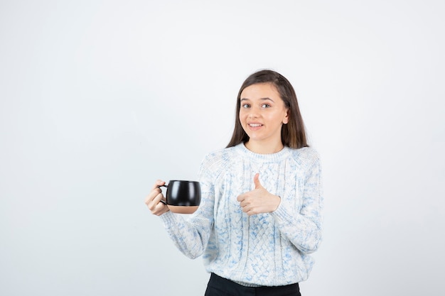 Jeune femme tenant une tasse de café chaud et montrant le pouce vers le haut