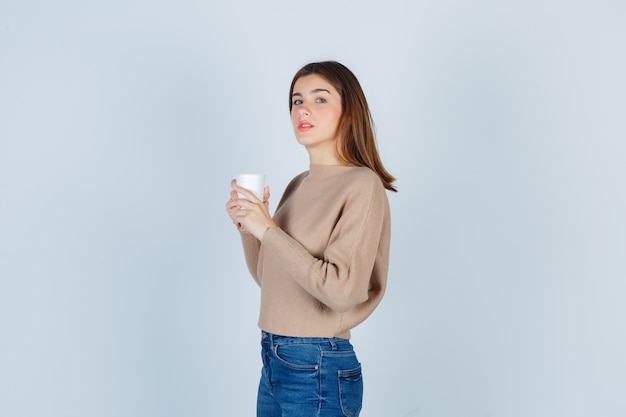 Jeune femme tenant une tasse de papier, à la recherche, debout sur le côté en pull, jeans et à la réflexion.