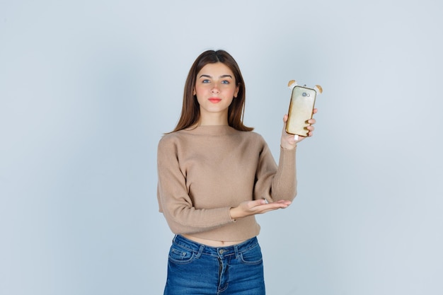 Jeune femme tenant un téléphone portable dans un pull beige, un jean et l'air confiant. vue de face.