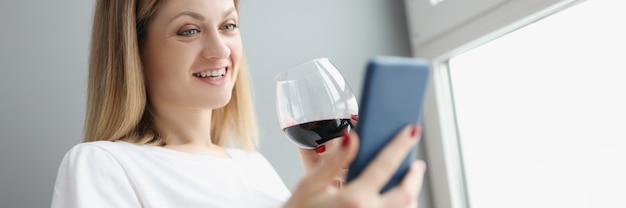 Jeune femme tenant un téléphone portable dans ses mains et buvant du vin rouge dans un verre à la maison