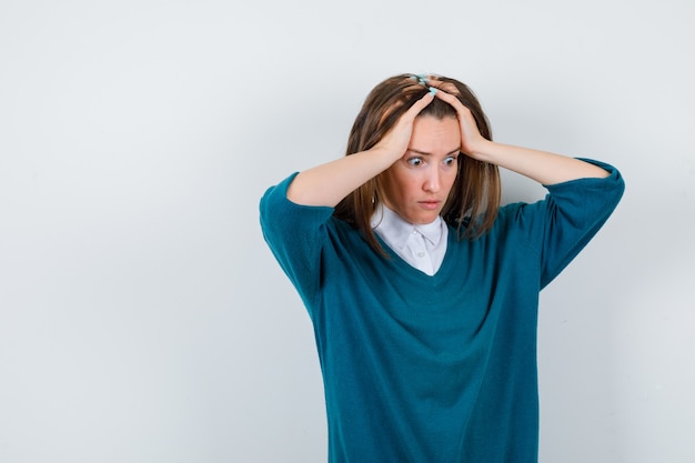 Jeune femme tenant la tête avec les mains, regardant vers le bas dans un pull sur une chemise et l'air perplexe, vue de face.