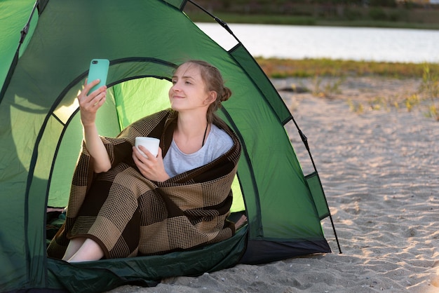 Jeune femme en tente buvant du café et prenant un selfie. Camping sur plage de sable.