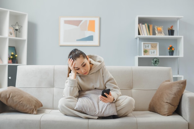 Jeune femme en tenue décontractée assise sur un canapé à l'intérieur de la maison à l'aide d'un smartphone regardant l'écran confus et triste