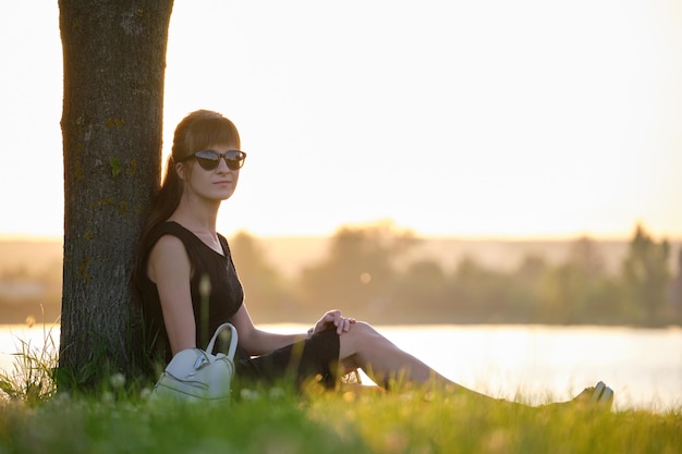 Jeune femme en tenue décontractée assise sur une pelouse d'herbe verte relaxante au bord du lac lors d'une soirée chaude Vacances d'été et concept de voyage