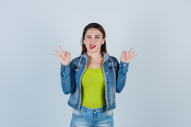 Jeune femme en tenue de denim montrant un geste correct tout en clignant des yeux, en tirant la langue et en ayant l'air enjouée, vue de face.
