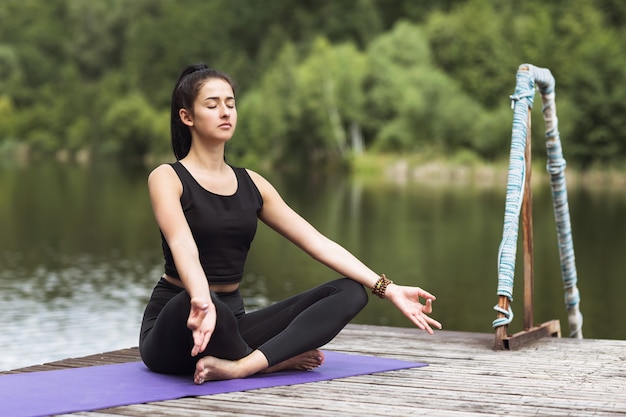 Une jeune femme en tenue de sport médite en position du lotus sur un pont en bois au bord de la rivière