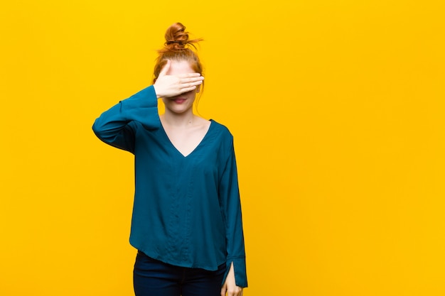 Jeune femme tête rouge couvrant les yeux avec une main, se sentant effrayé ou anxieux, se demandant ou attendant aveuglément une surprise contre le mur orange