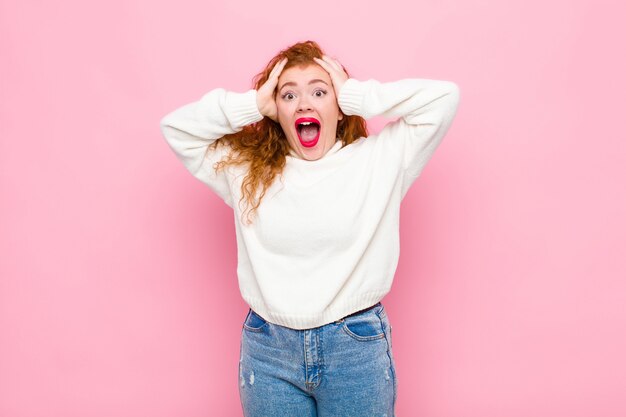 Jeune femme tête rouge levant les mains à la tête, bouche ouverte, se sentant extrêmement chanceux, surpris, excité et heureux sur le mur rose