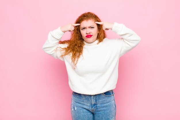 Jeune femme à tête rouge avec un regard sérieux et concentré, remue-méninges et penser à un problème difficile contre le mur rose