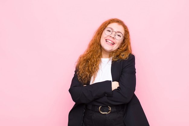 Jeune femme tête rouge rire joyeusement avec les bras croisés, avec une pose détendue, positive et satisfaite contre le mur rose