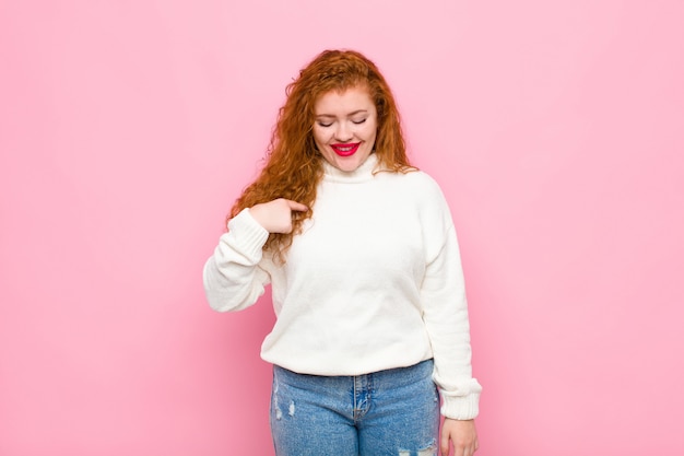 Jeune femme tête rouge souriant joyeusement et nonchalamment, regardant vers le bas et pointant vers la poitrine contre le mur rose