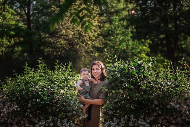 Photo une jeune femme tient un petit bébé dans ses bras. une belle mère se promène avec sa fille dans un parc verdoyant près des buissons de jasmin