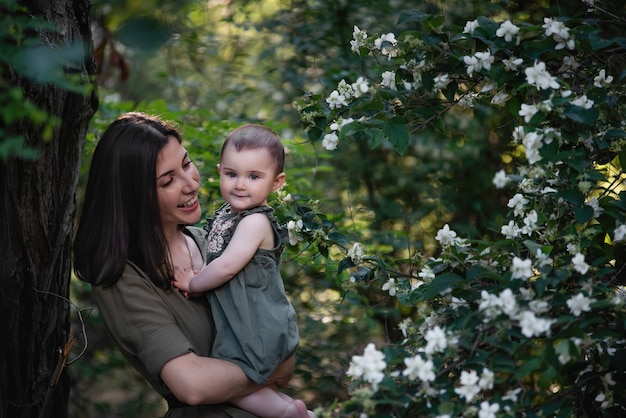 Une jeune femme tient un petit bébé dans ses bras. Une belle mère se promène avec sa fille dans un parc verdoyant près des buissons de jasmin