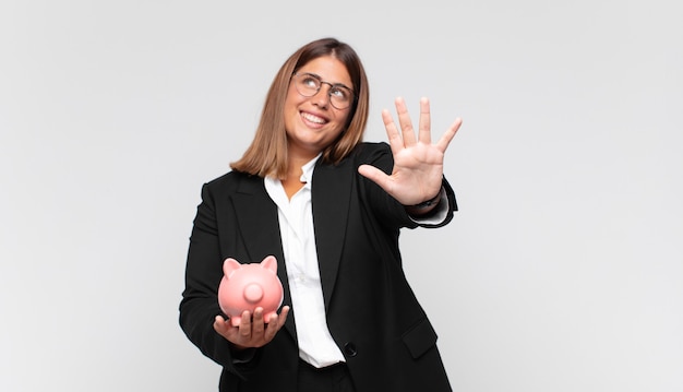 Jeune femme avec une tirelire souriant et à la convivialité, montrant le numéro cinq ou cinquième avec la main en avant, compte à rebours