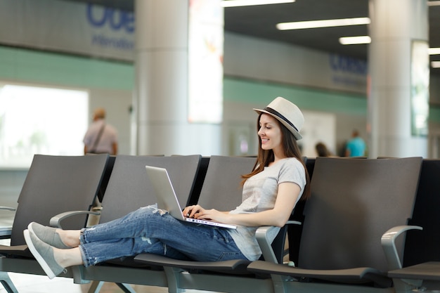 Jeune femme touriste calme au chapeau travaillant sur un ordinateur portable en attendant dans le hall de l'aéroport international